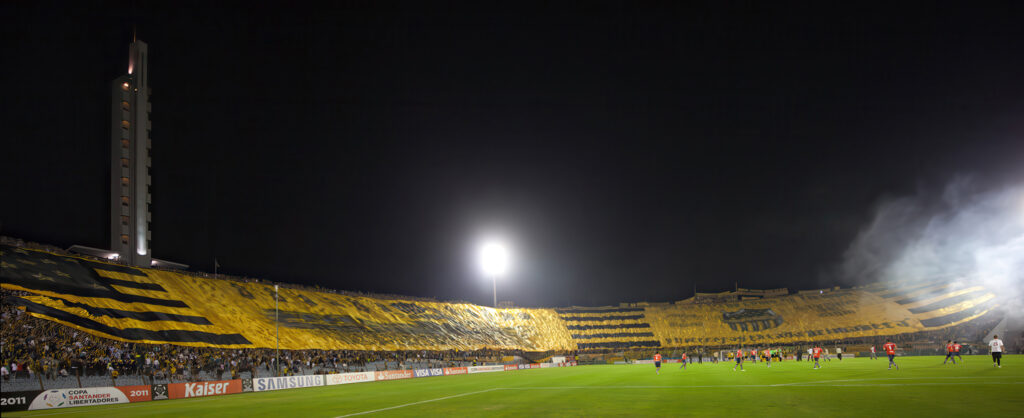 Le Peñarol détient le record du plus grand drapeau avec 309 m de large. Ici, lors de sa première présentation, le 12 avril 2011 © Jimmy Baikovicius https://www.flickr.com/photos/jikatu/5615126082/. On peut y voir une locomotive rappelant l’origine du club. La bannière normale reprend le graphisme du drapeau uruguayen avec ses lignes horizontales, mais le soleil est remplacé par des étoiles. Le résultat est graphiquement proche de la bannière des USA, sauf les couleurs l’or et le noir. Cette bannière a été utilisée pour la première fois pour une rencontre contre Independiente (cité en début de cet article…)